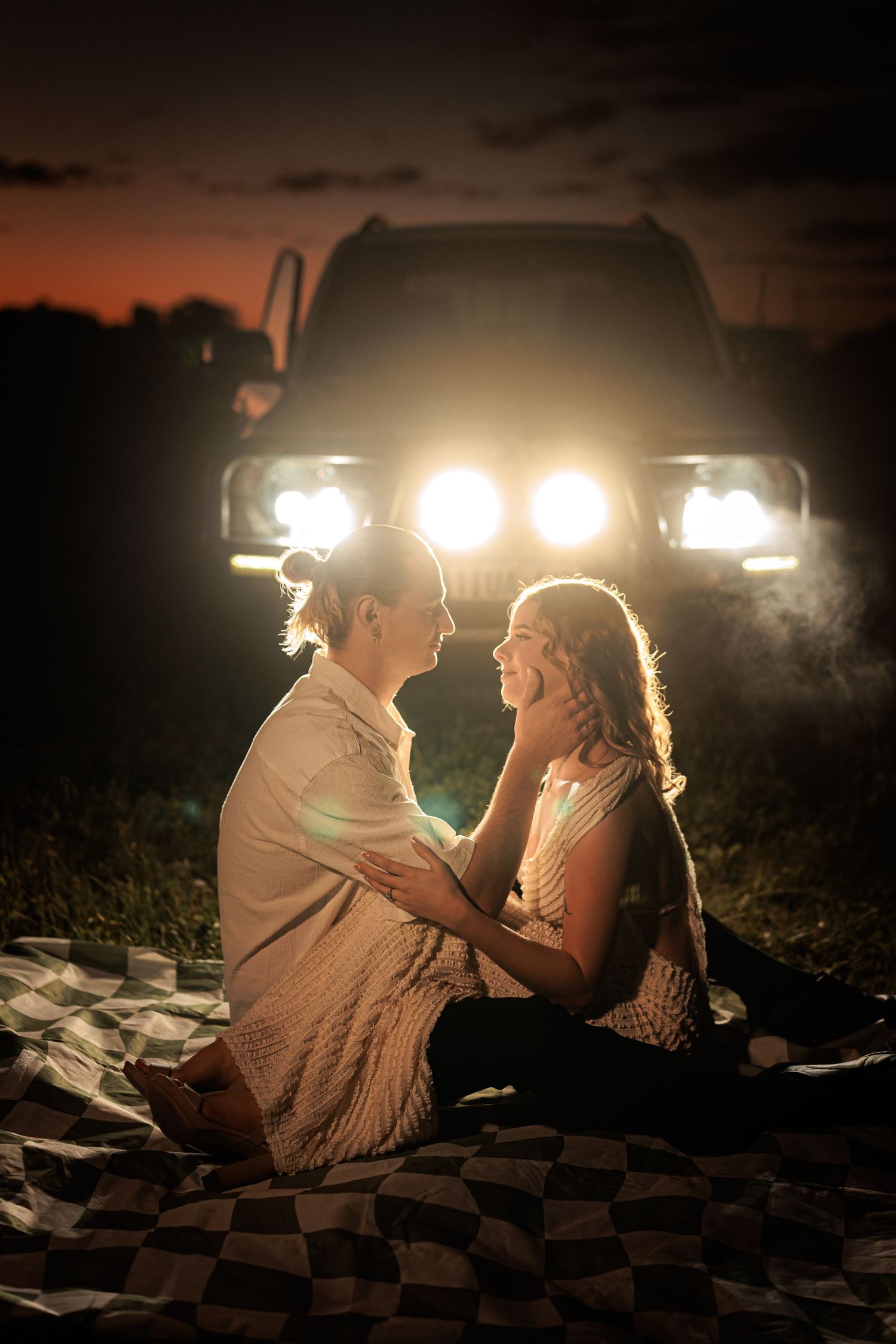 Two women sit facing each other on a blanket at night, illuminated by car headlights against a sunset sky.