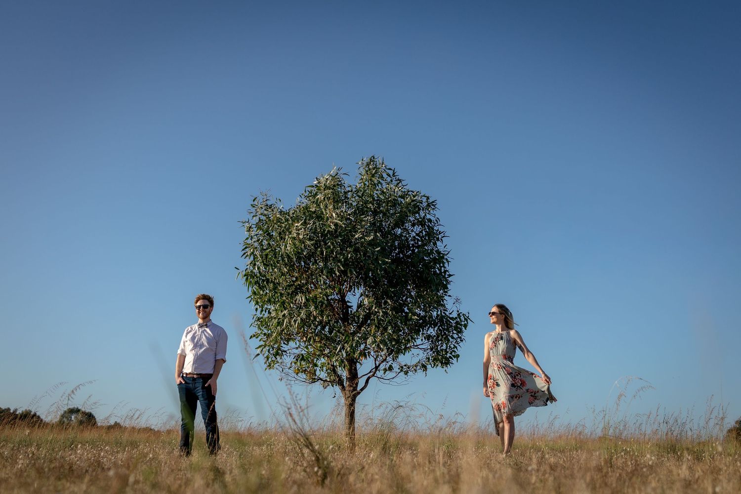 Couple stands apart on either side of lone tree in open field under blue sky.
