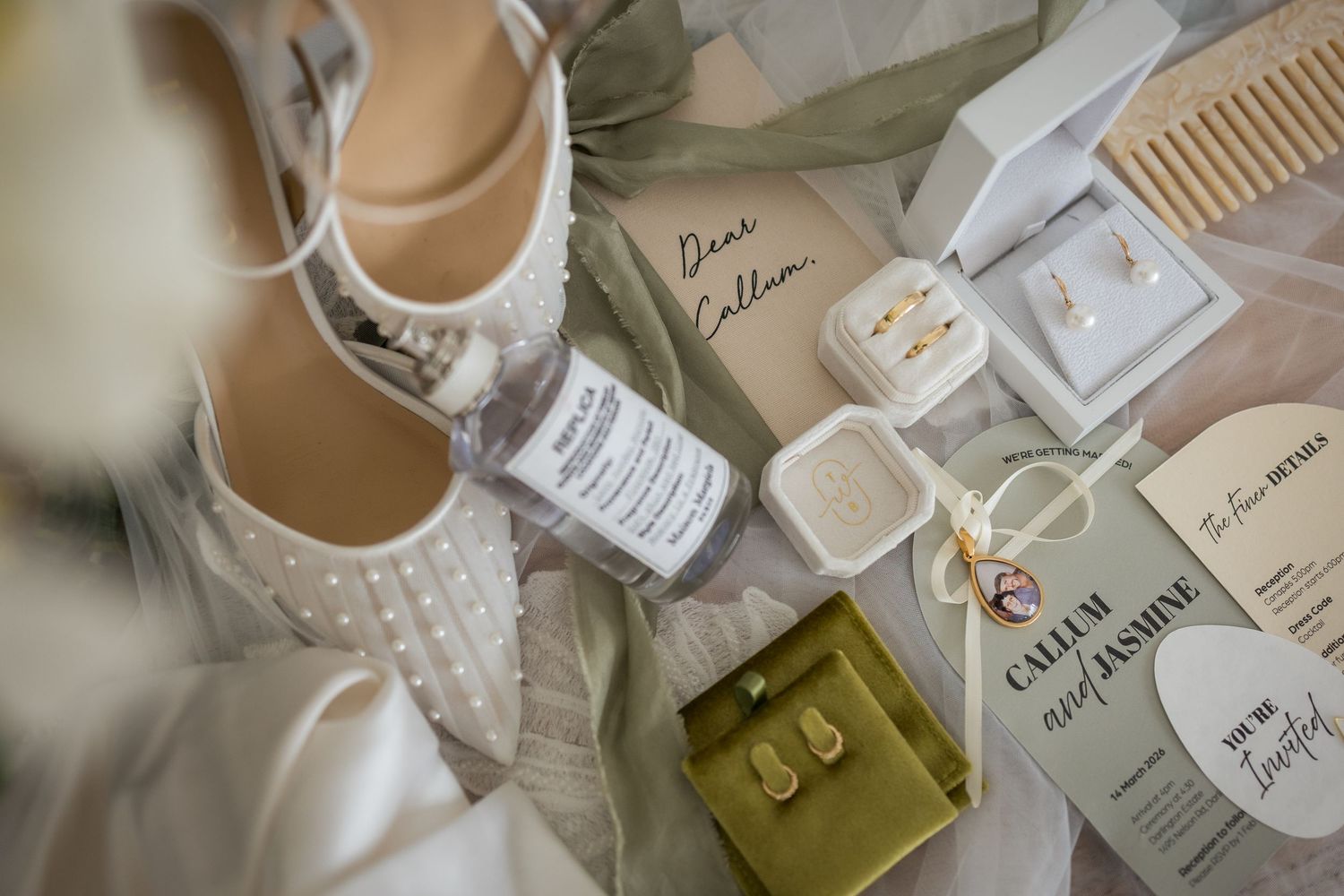 Bridal flat lay with pearl shoes, gold earrings, wooden comb, and personalized wedding stationery on white fabric.