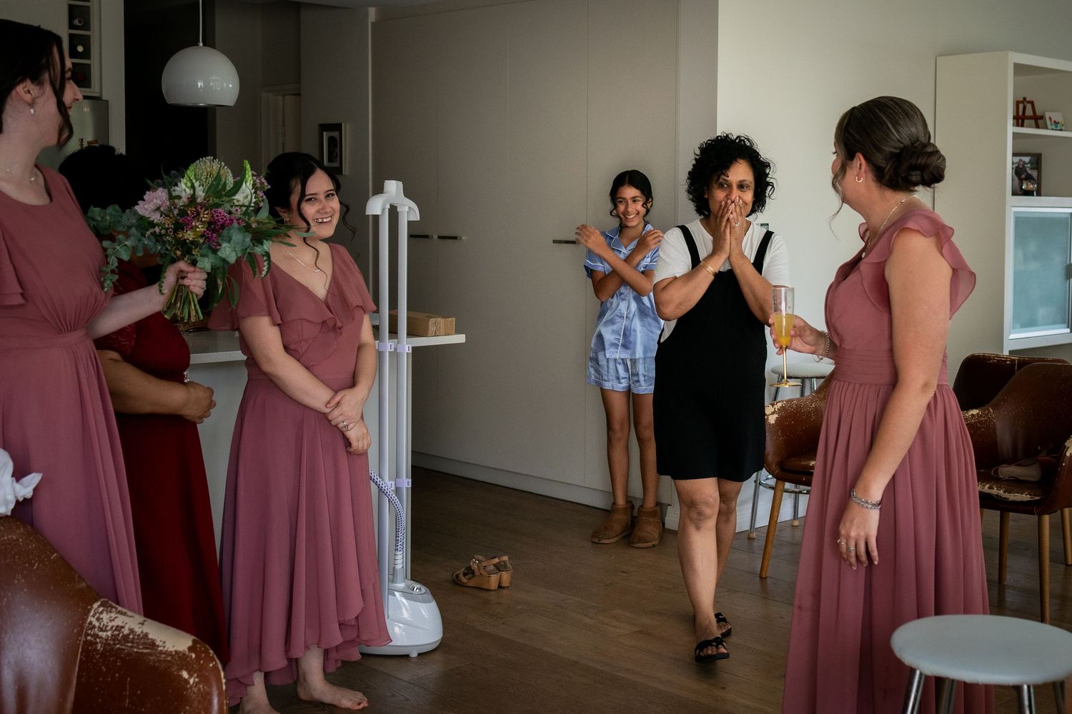Bridesmaids in dusty rose dresses gather as emotional woman clasps hands in reaction during wedding preparations indoors.