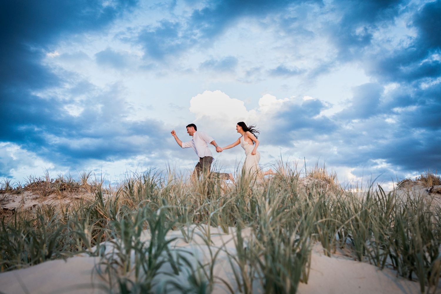 Couple holding hands dancing on sandy dunes with lush grass and dramatic cloudy sky backdrop.