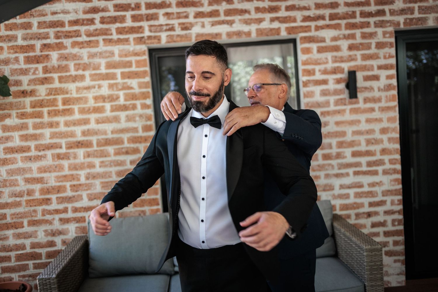 Groom in black tuxedo gets jacket adjusted by older man in suit near brick wall.