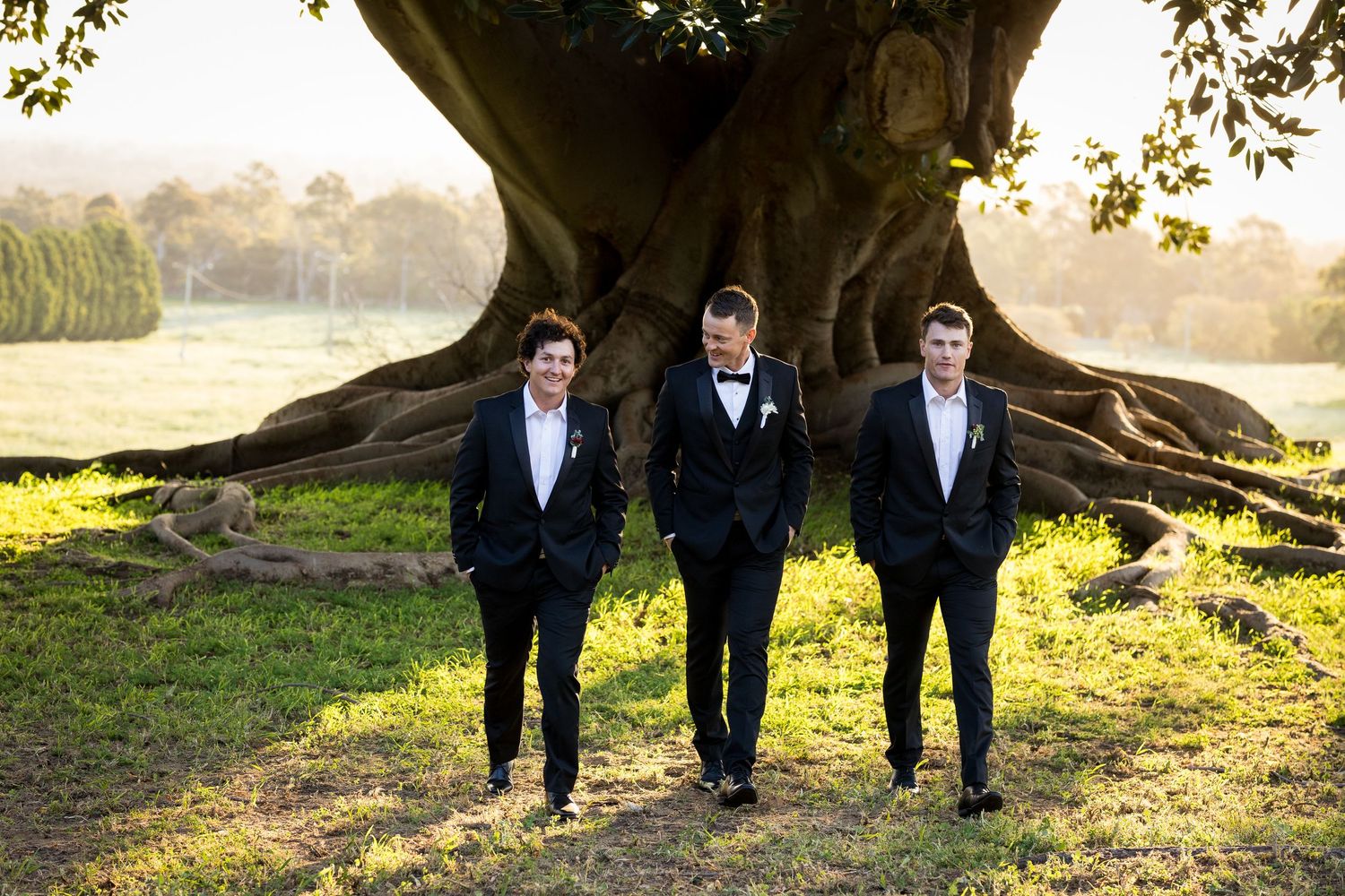 Wedding party members walking through park with golden sunlight filtering through trees.