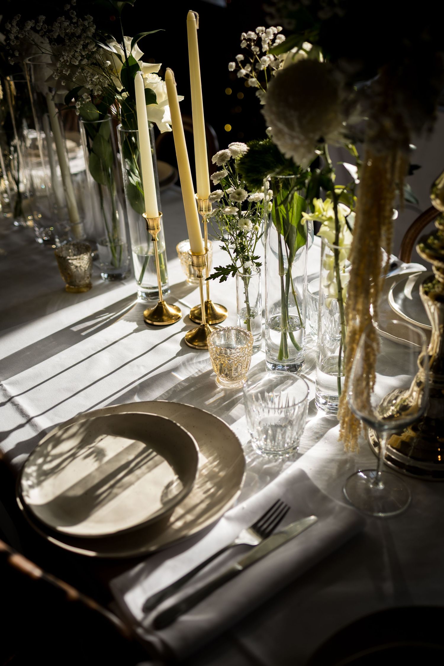 Elegant wedding table setting with white flowers, gold candlesticks, crystal glasses, and a white plate on a linen tablecloth.