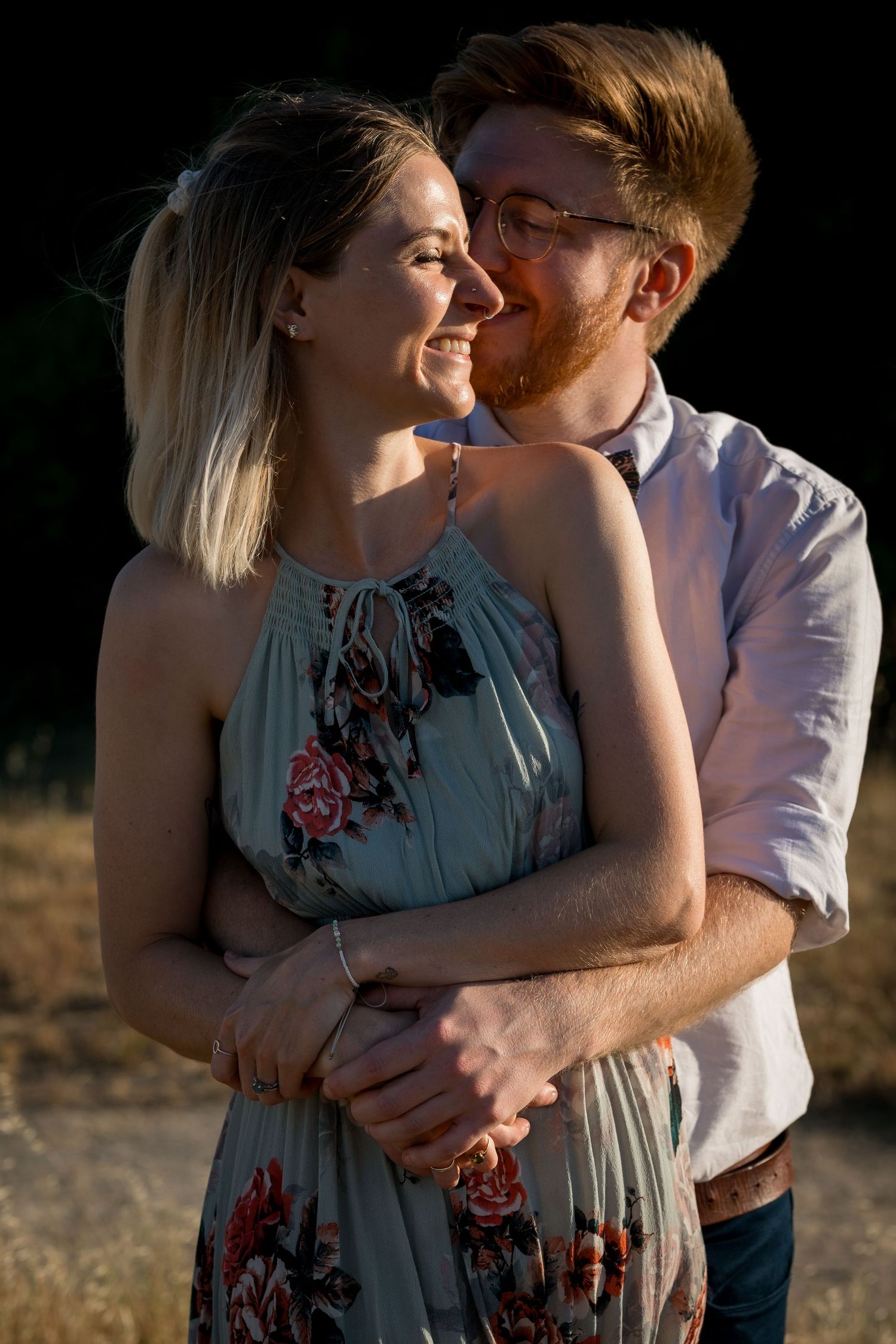 A smiling couple embraces outdoors at golden hour, woman in floral dress, man with red beard and glasses.