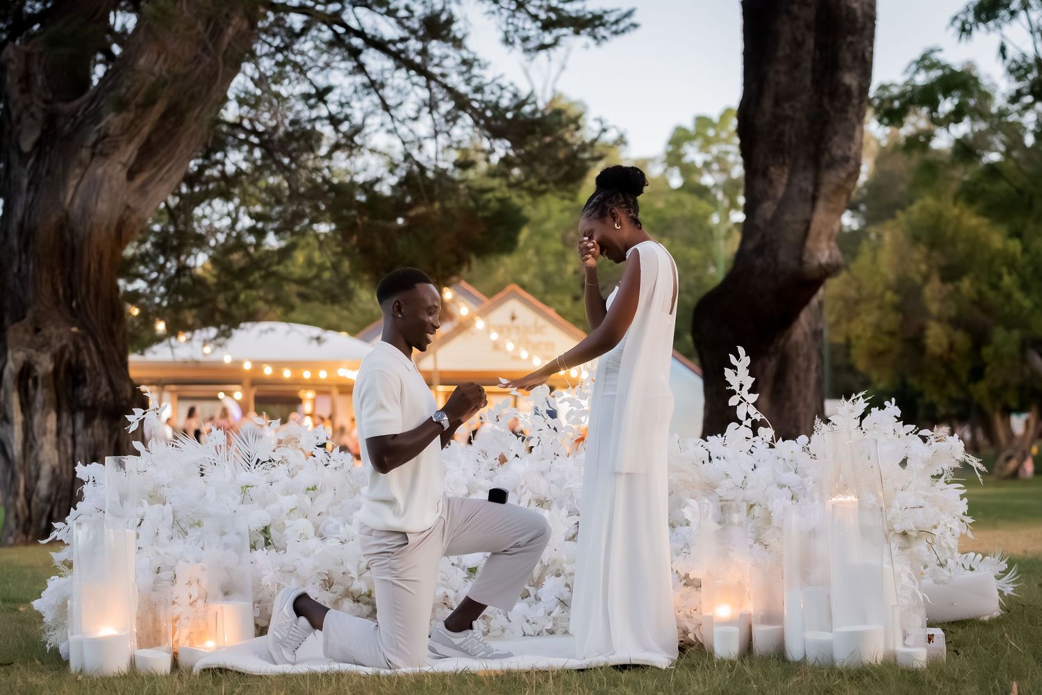 Man proposes to woman on one knee surrounded by white flowers and candles in romantic outdoor setting.