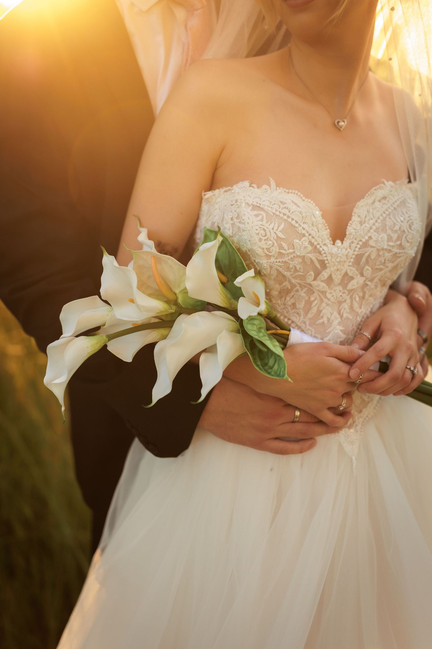 Bride in strapless lace ballgown holding white calla lily bouquet, embraced by groom in golden sunset light.