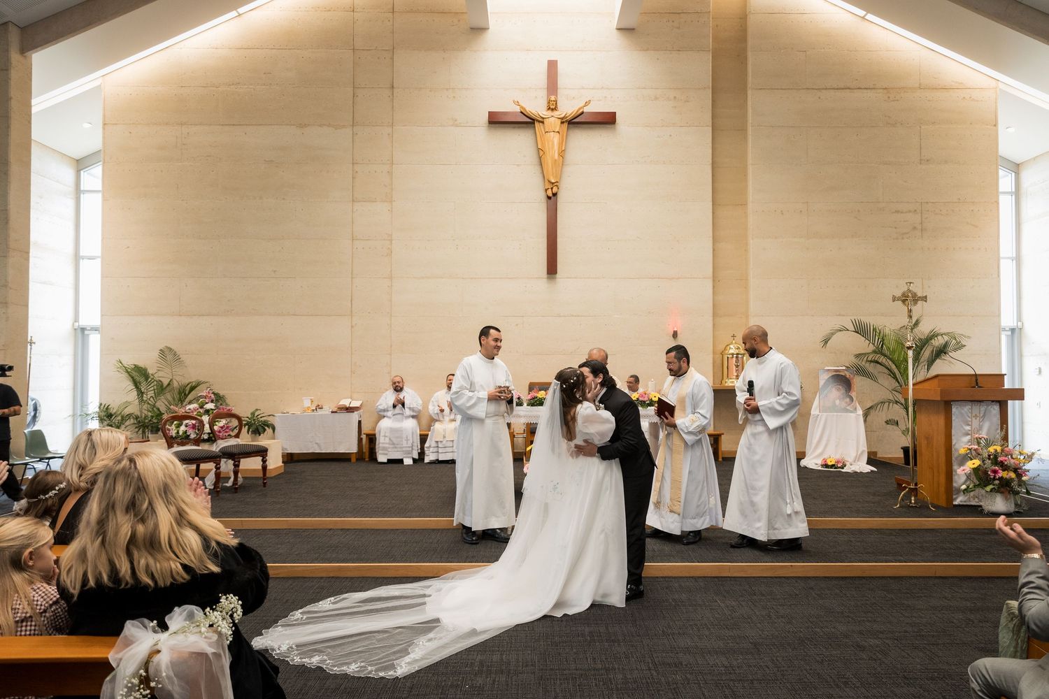 Bride and groom share first kiss at Catholic church wedding ceremony with altar servers and cross backdrop.