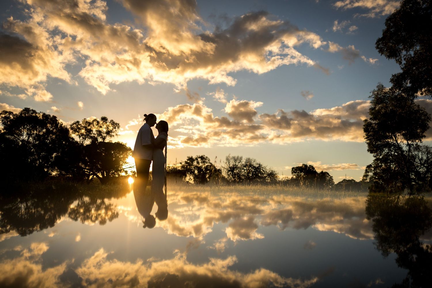 Couple silhouetted against golden sunset reflected in still water, surrounded by trees and dramatic clouds. at Guildford, Perth