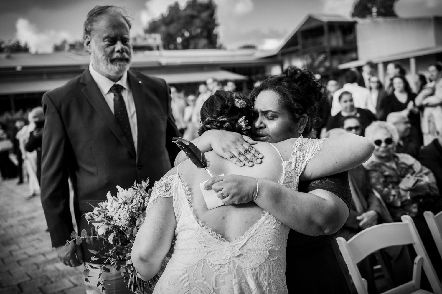 Black and white wedding photo of bride embracing a guest emotionally while a man in suit watches during outdoor ceremony.