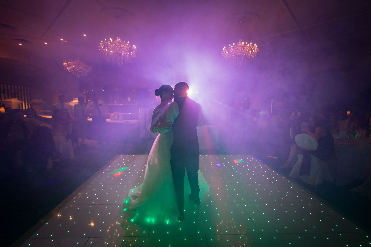 Couple sharing first dance on illuminated LED dance floor with purple lighting and elegant chandeliers at wedding reception.