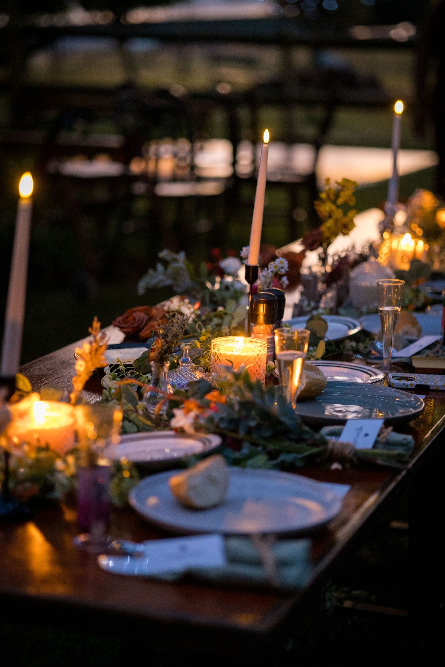 Elegant candlelit outdoor wedding reception table adorned with floral arrangements, tapered candles, and fine dinnerware at dusk.