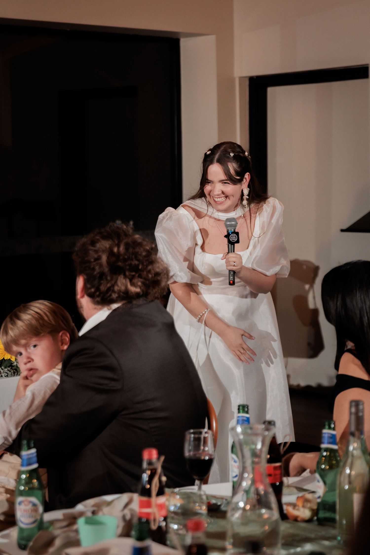 Bride in white dress giving a speech with microphone at wedding reception dinner.