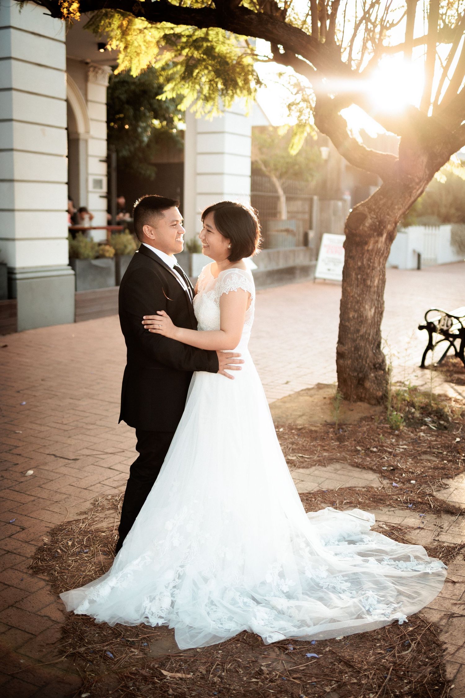 A bride and groom embrace outdoors in golden sunset light, she in a white lace gown, he in a black tuxedo.