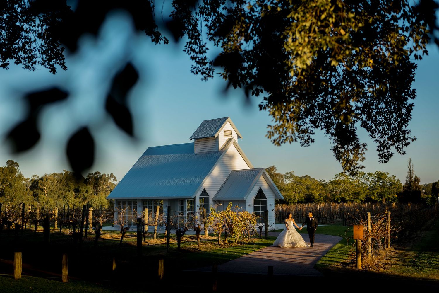 Bride and groom stand before a charming white chapel surrounded by lush greenery and vineyards at golden hour.