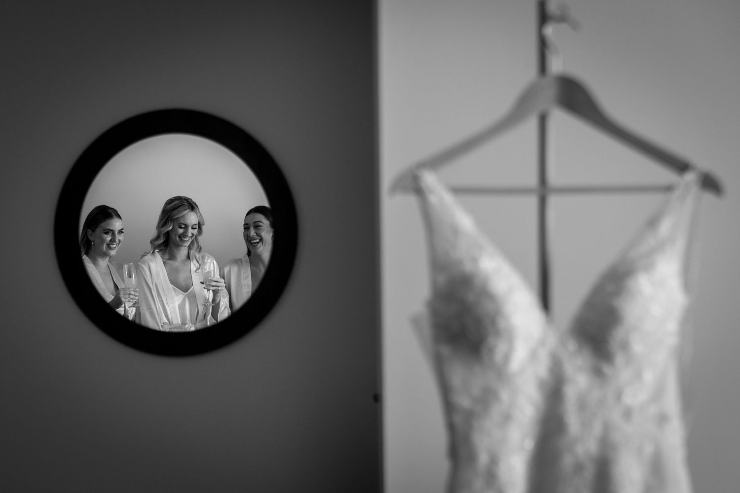 Black and white wedding photo of bride reflected in round mirror with bridesmaids, lace gown hanging in foreground.