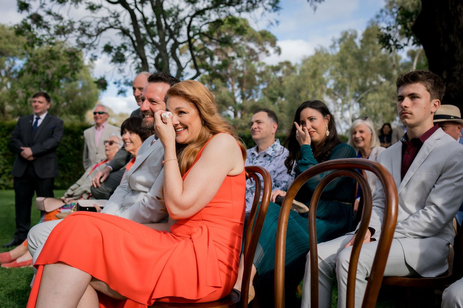 Wedding party members in coral and blue dresses gather outdoors under trees.