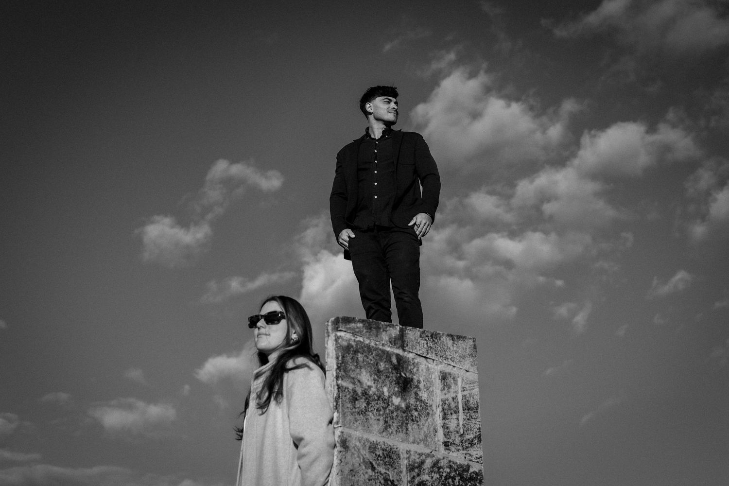 Black and white artistic portrait shot with dramatic clouds and architectural elements.