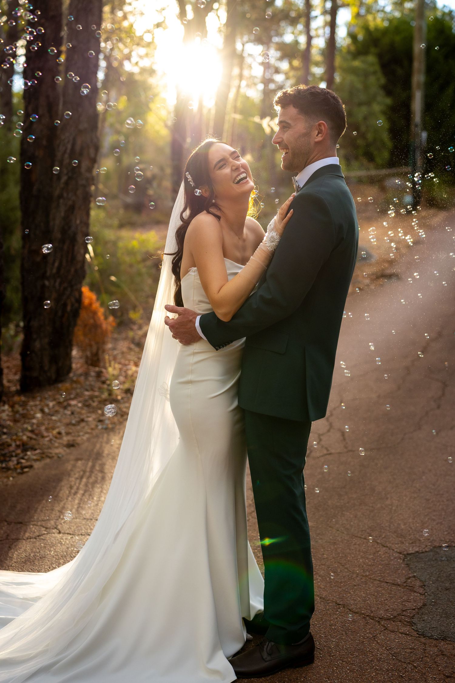Bride and groom embracing outdoors in golden sunlight with bubbles floating around them, bride in white gown laughing joyfully.
