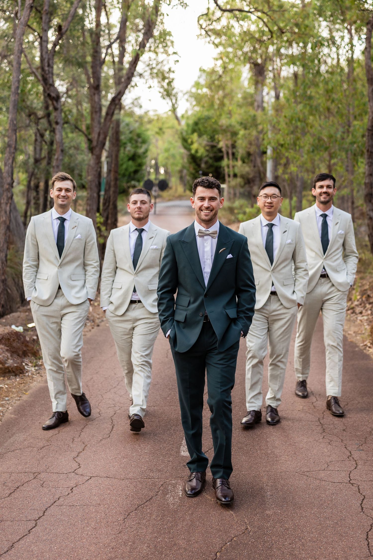 Groom in green suit leads four groomsmen in beige suits walking down a tree-lined path.