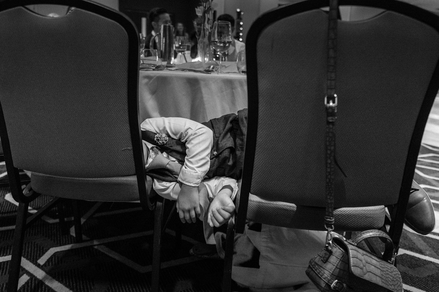 Black and white photo of a child sleeping curled up between banquet chairs at a formal event.