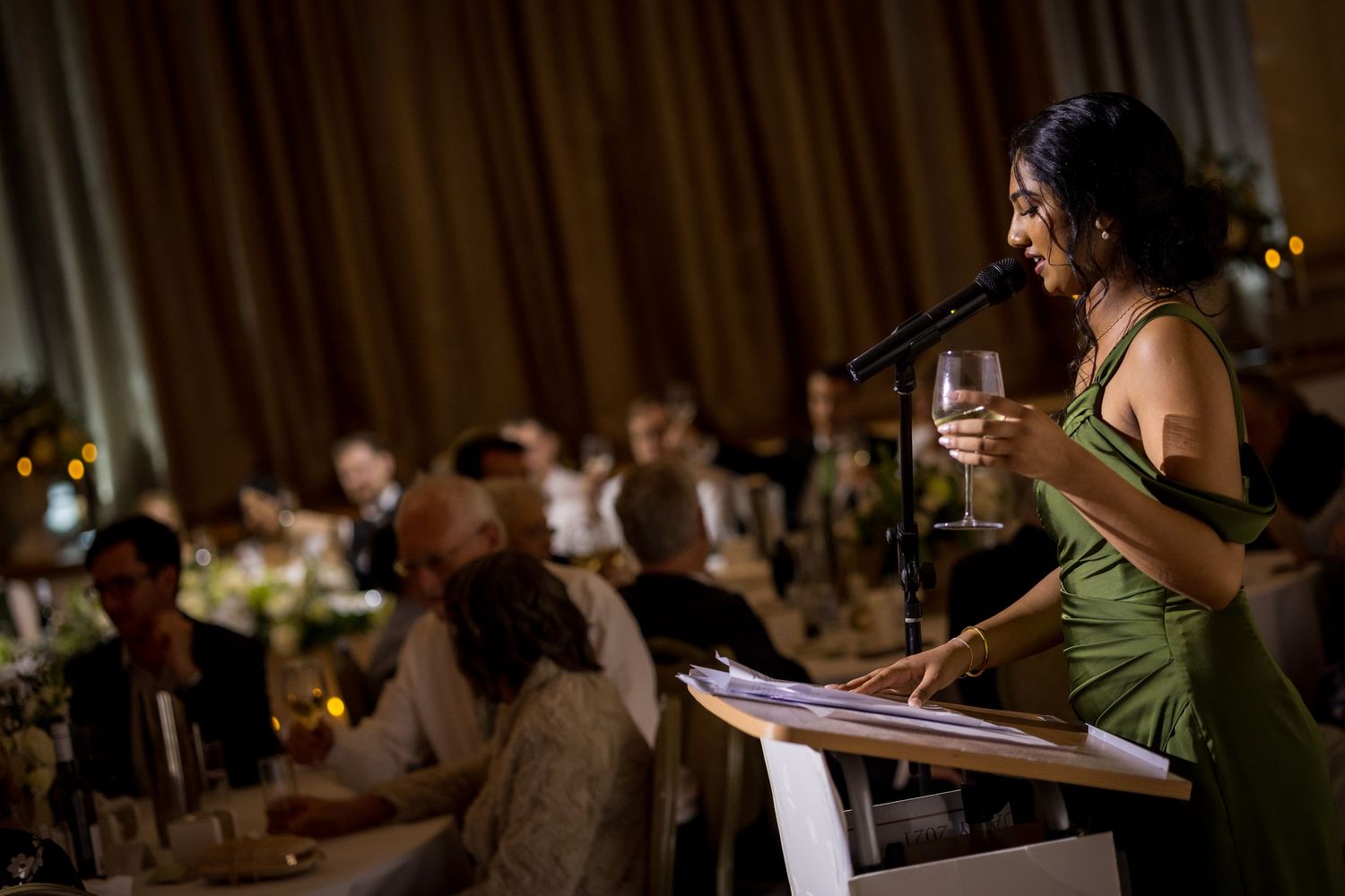 Woman in green dress gives toast at microphone holding wine glass during elegant wedding reception dinner. at Montgomery Hall