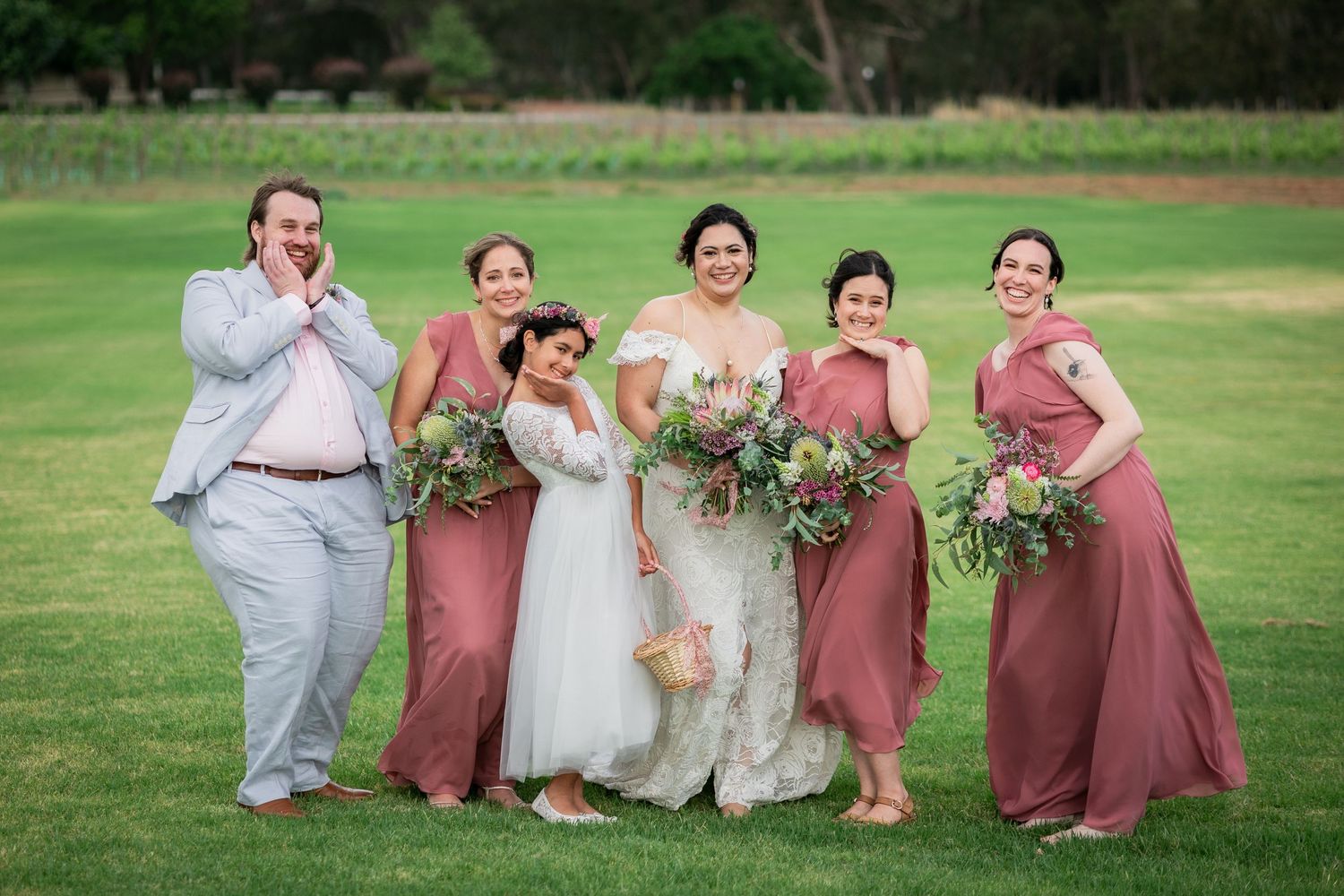 Bride poses with bridesmaids in dusty rose gowns and groomsman on lush green lawn.