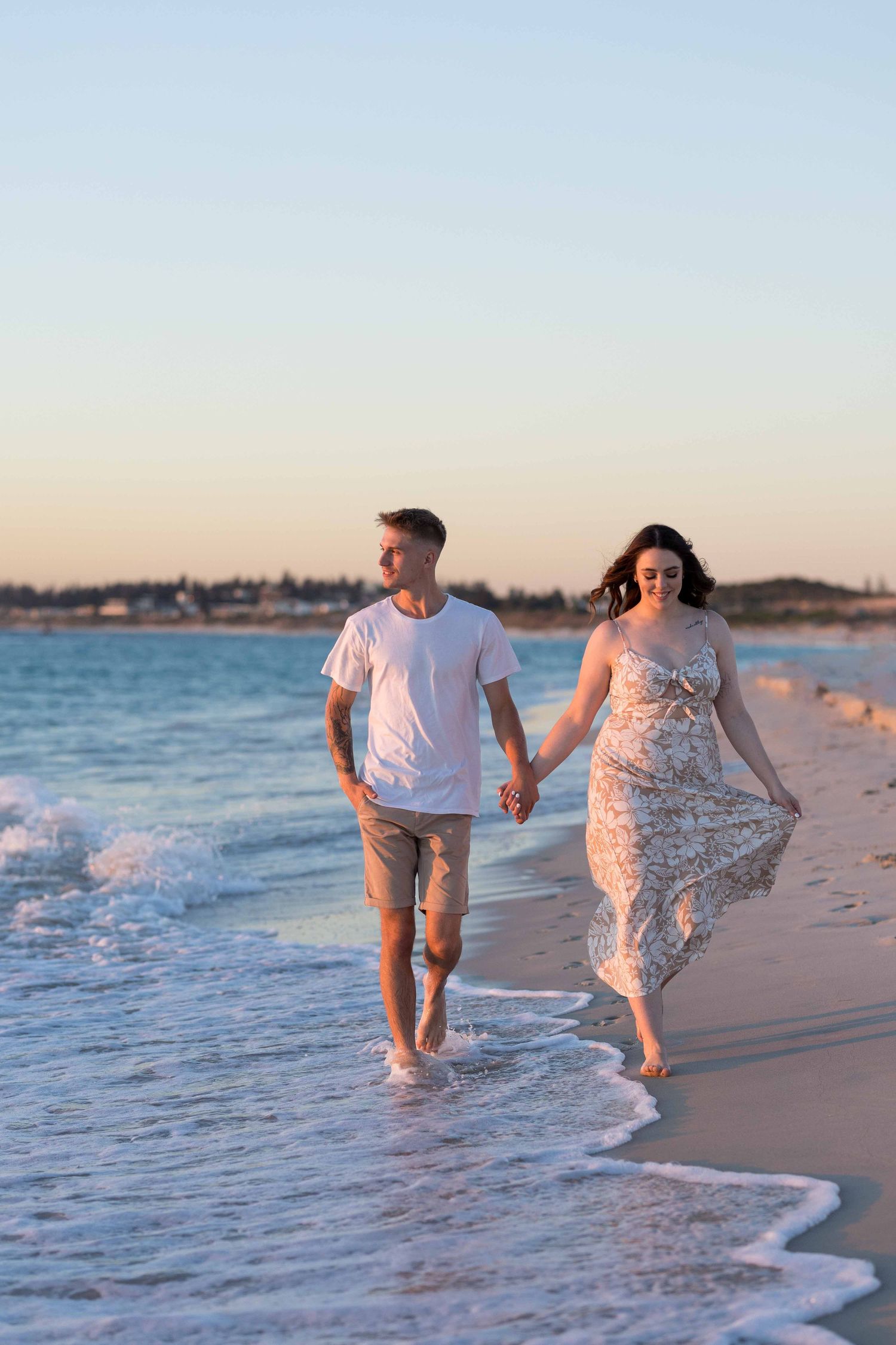 Couple holding hands walking along a sandy beach at sunset, man in white tee and shorts, woman in floral dress.
