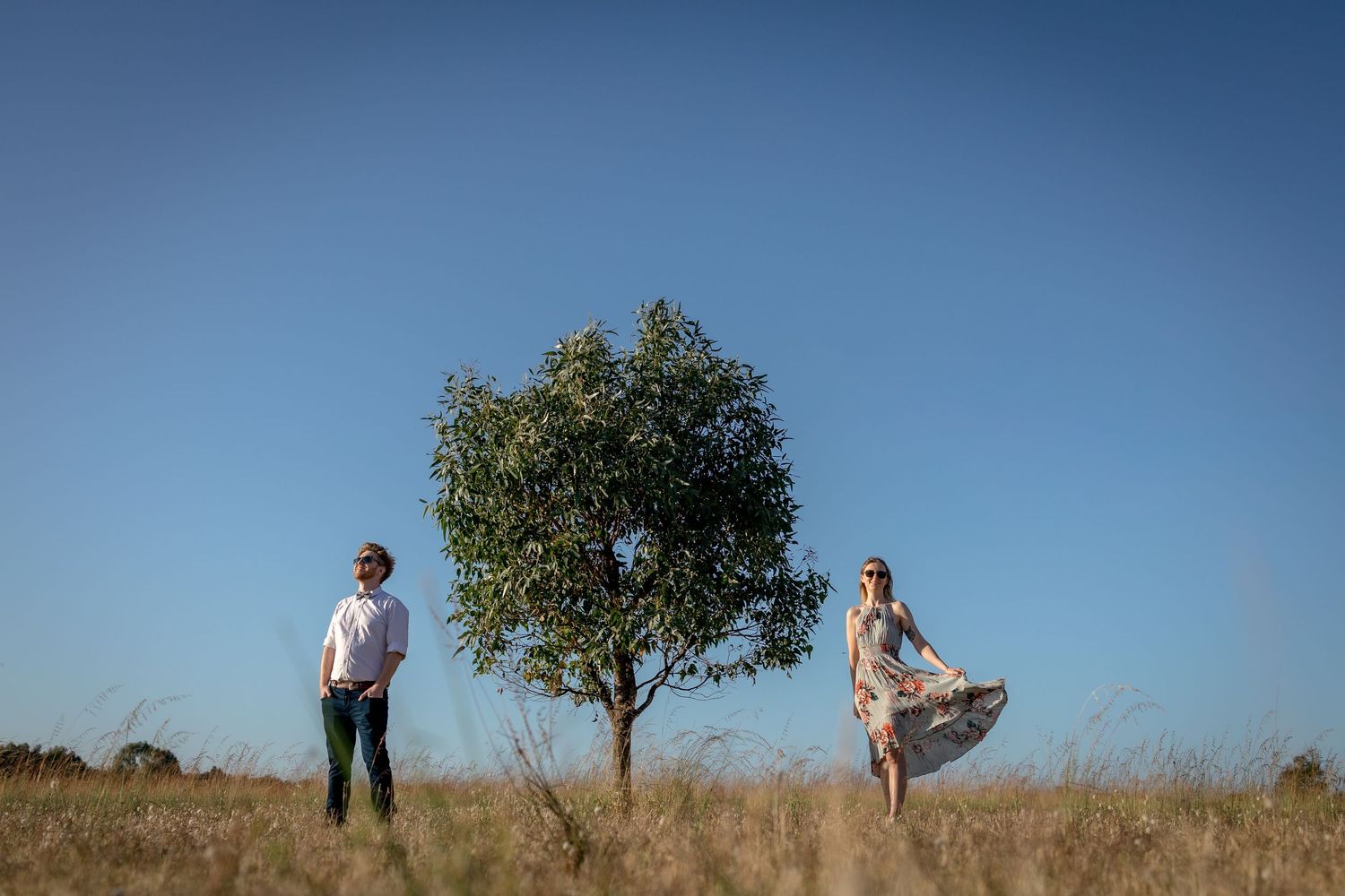 A couple stands together in a scenic field with blue sky and trees in the background.