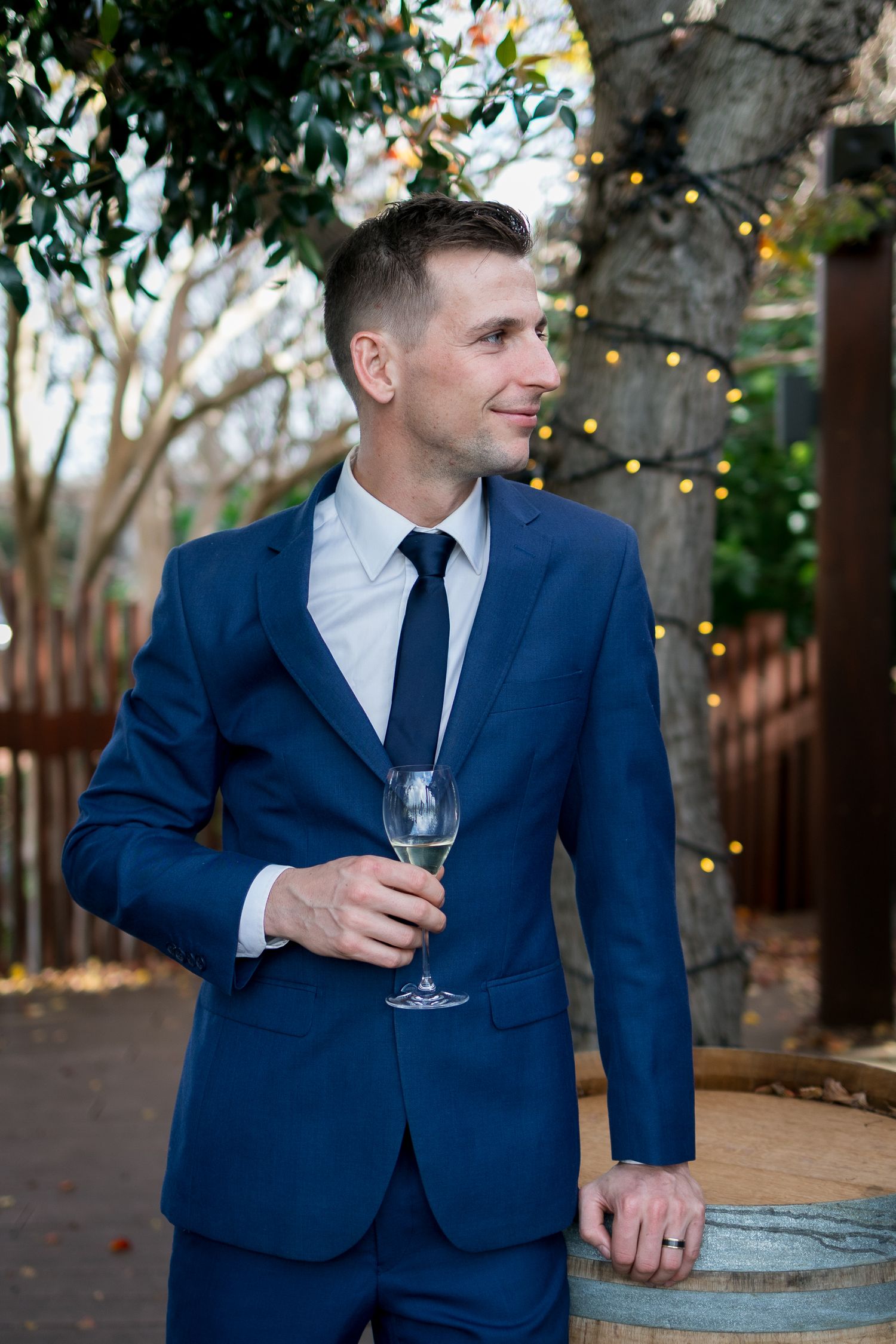 Person in navy blue suit holds champagne glass near outdoor wine barrels.