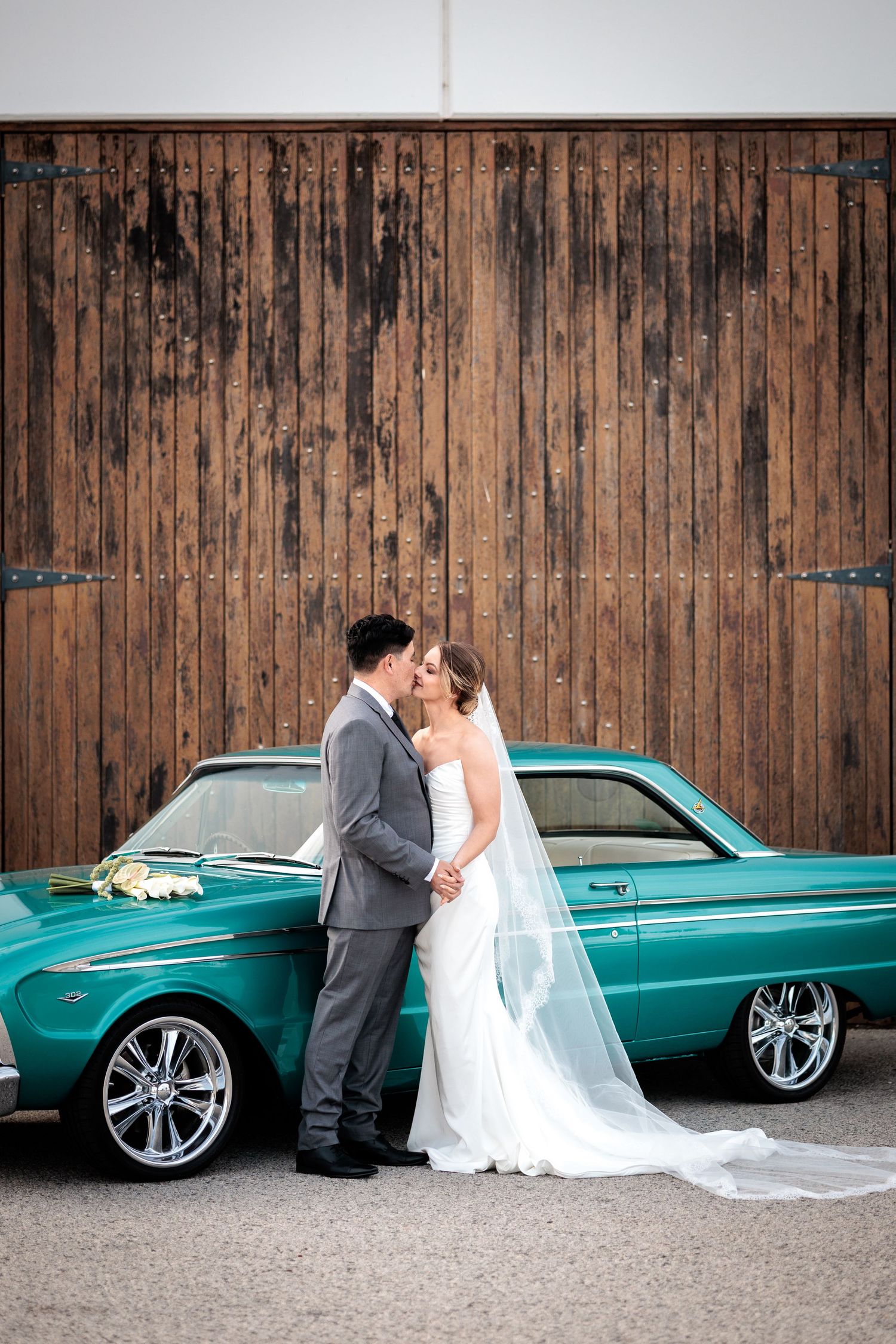 Bride and groom kissing in front of a vintage teal car and rustic wooden barn wall.
