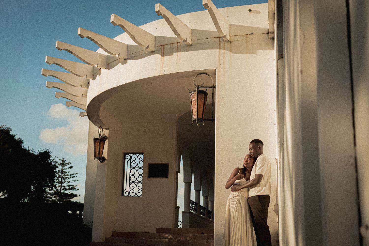 Couple embracing at golden hour beside a curved white building with a modern slatted roof, warm hanging lanterns and blue sky.