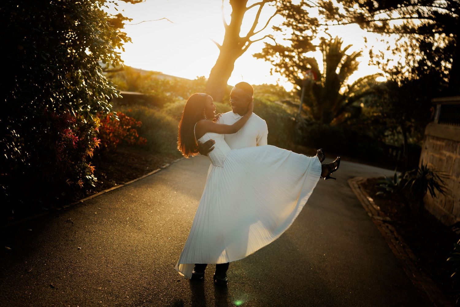 Groom lifting and spinning his bride in a flowing white wedding dress on a tree-lined path at golden hour sunset.