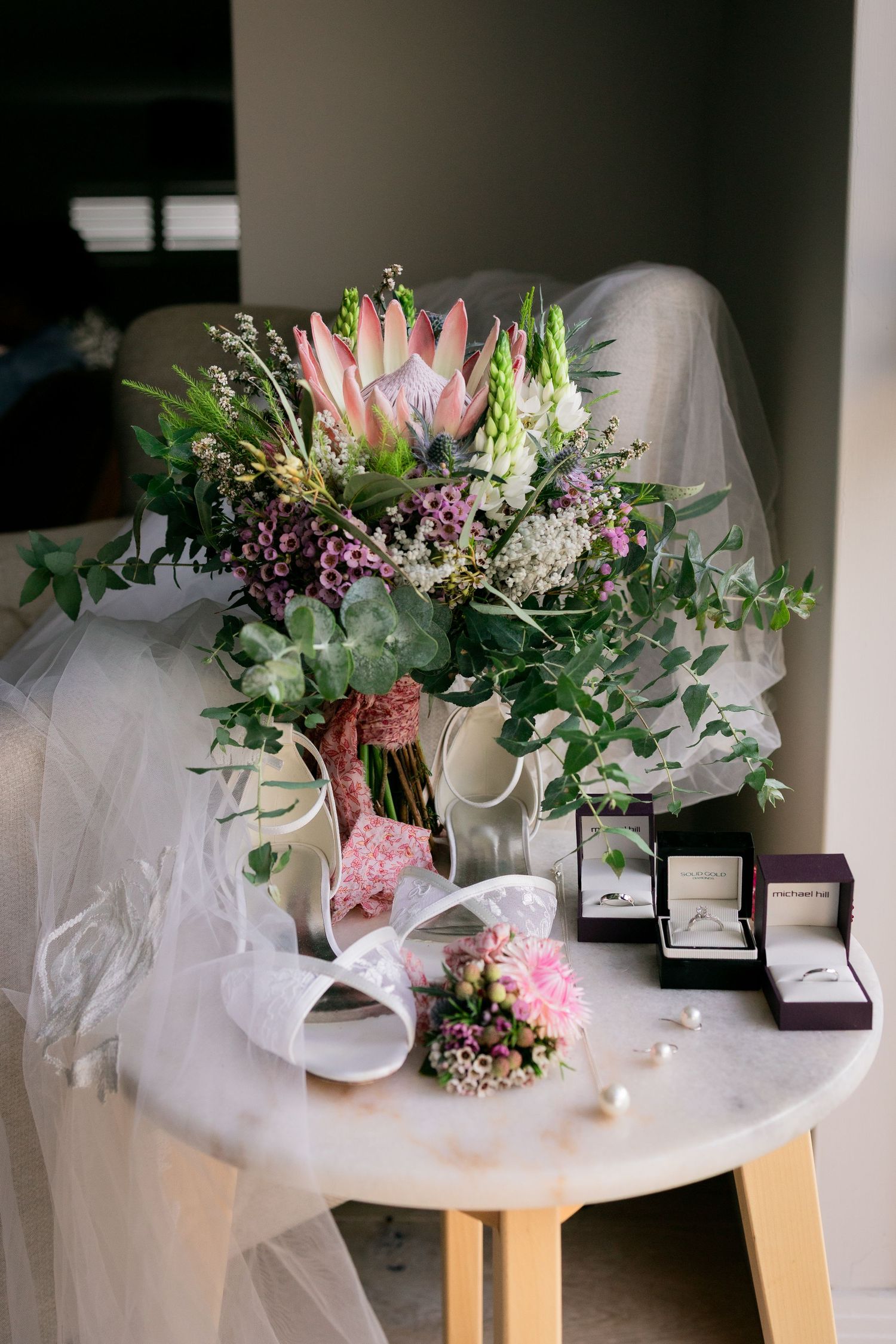 Lush bridal bouquet with protea and greenery beside ring boxes and veil on marble table.
