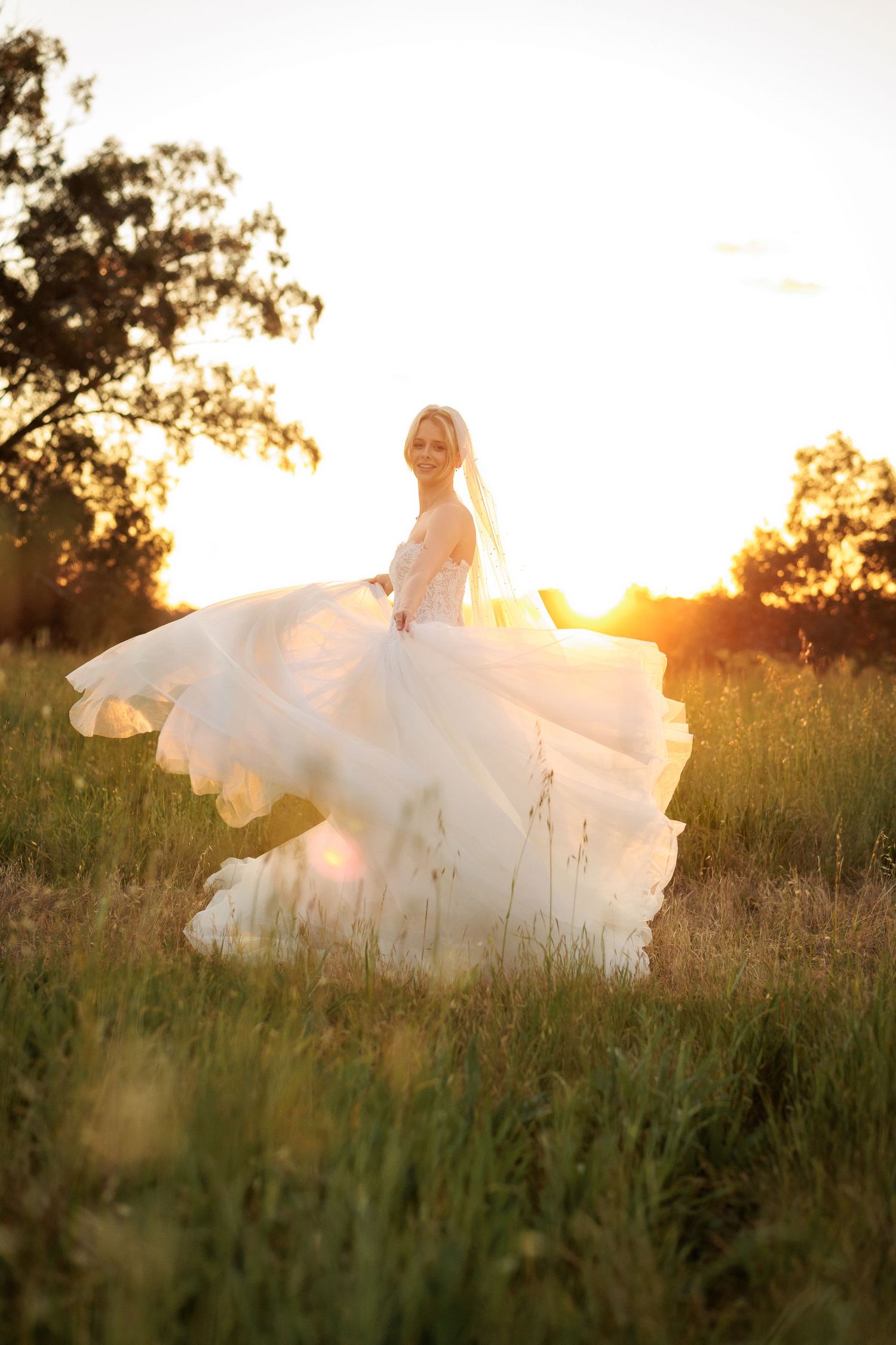 Bride in flowing white gown twirling in golden sunset field, veil and ruffled skirt billowing in warm evening light.