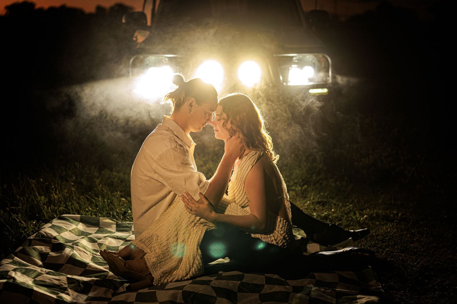 Romantic couple sitting on a blanket at dusk, illuminated by car headlights in a dreamy outdoor setting.