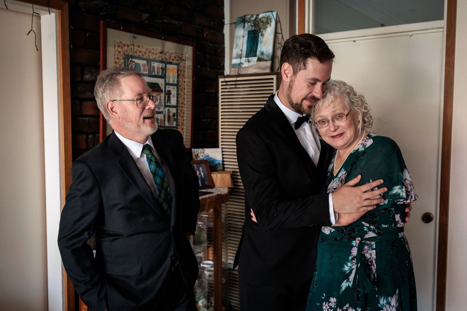 Groom in tuxedo embraces older woman in green floral dress while older man in suit smiles at wedding celebration.