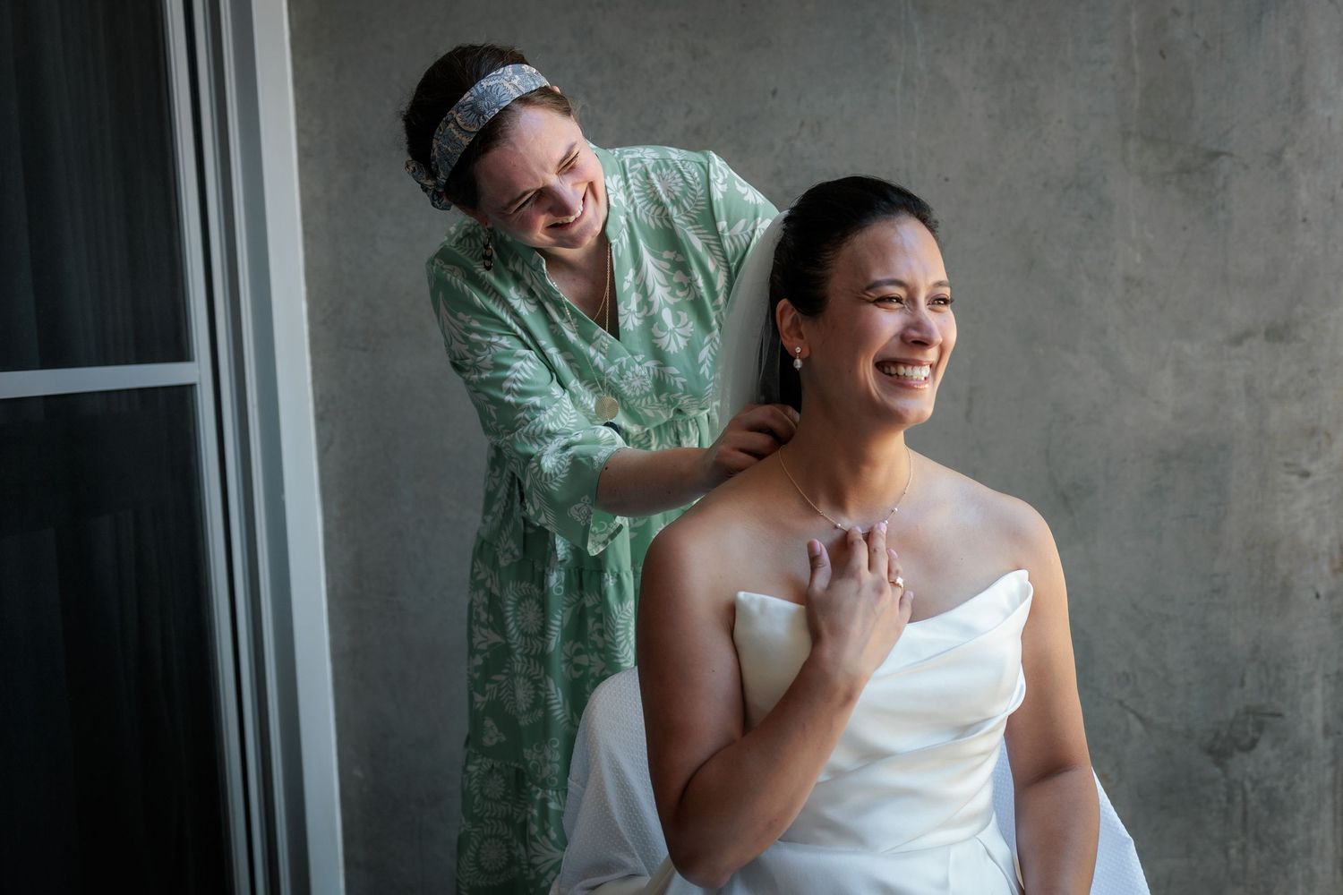 Bridesmaid in green robe helping bride in white strapless gown with necklace, both smiling on wedding day.