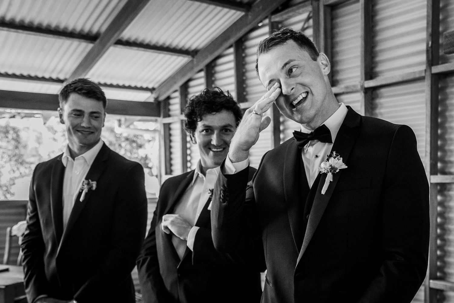 Three groomsmen in black tuxedos laughing and sharing a joyful moment together before a wedding ceremony.