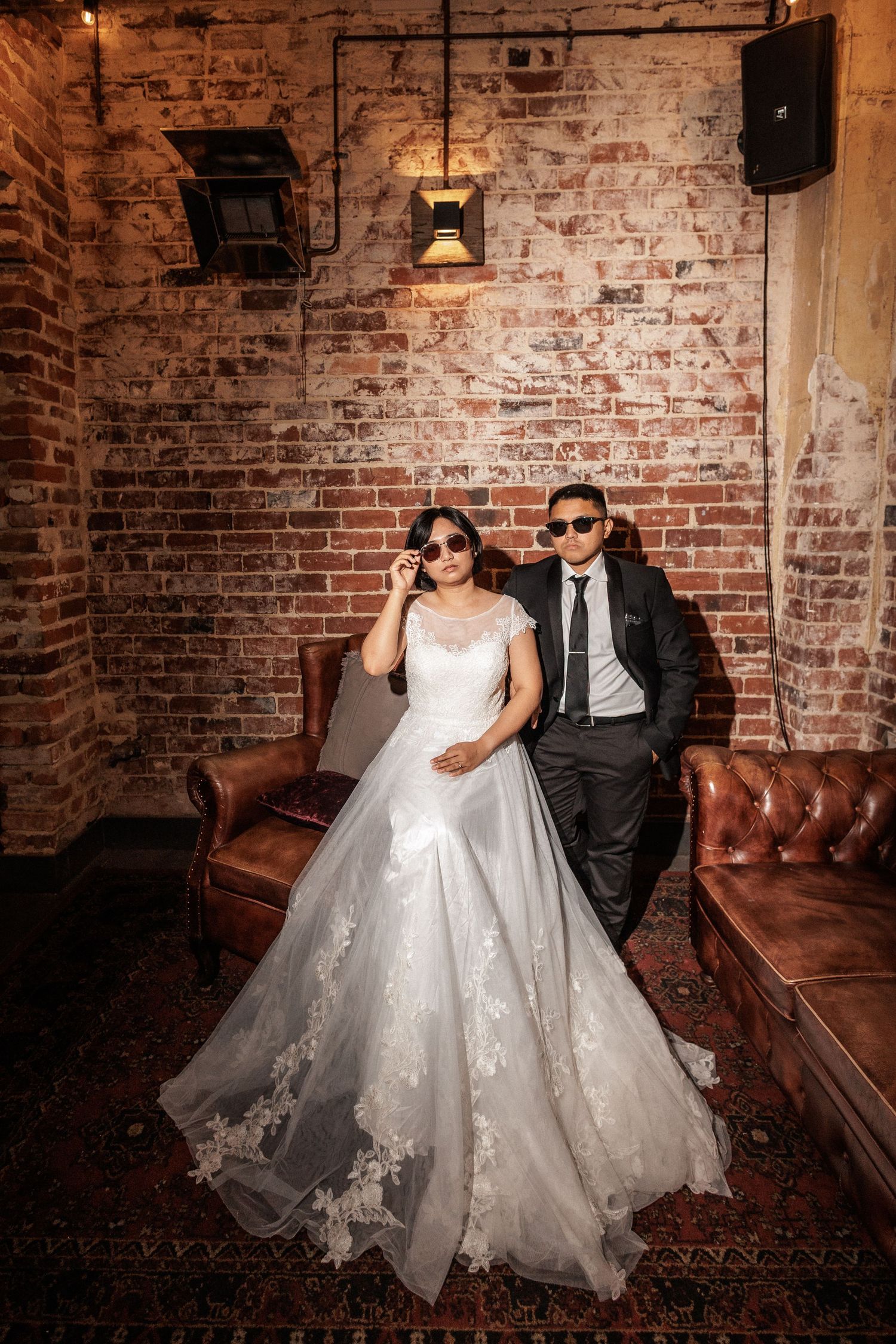 Bride in white lace gown and groom in black suit wearing sunglasses, posing on leather sofa against rustic brick wall.