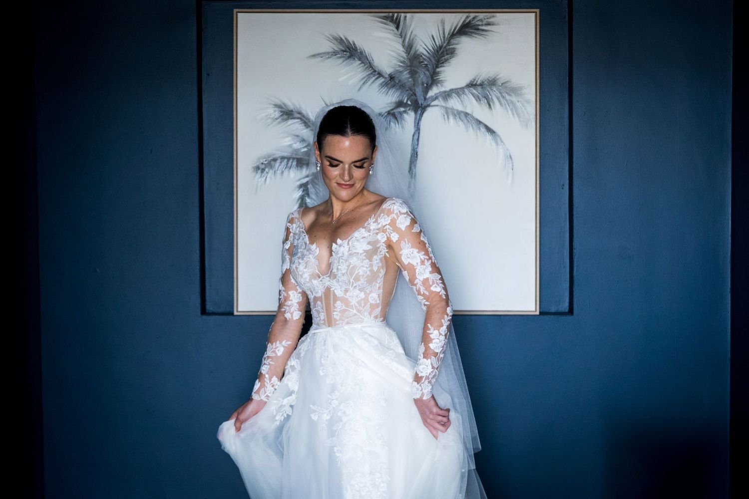 Bride in a long-sleeve lace wedding gown posing in front of a blue wall with palm art.