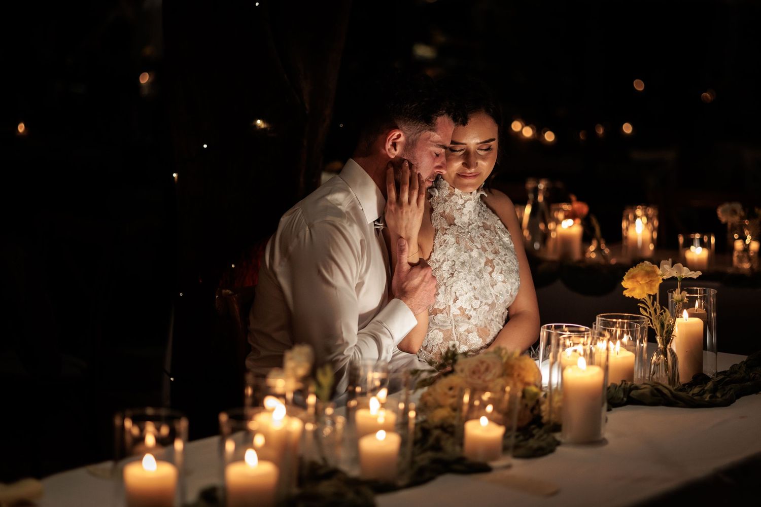 Romantic couple sharing an intimate moment at candlelit wedding reception table at night.