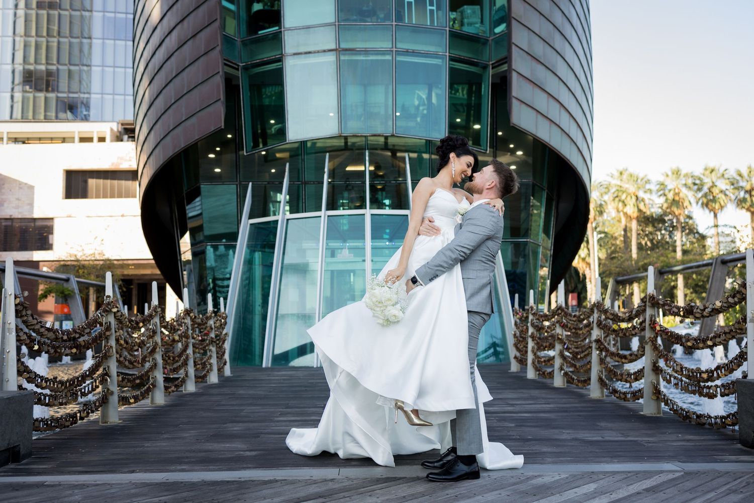 Groom lifting bride in white gown on decorated bridge outside modern glass building at outdoor wedding venue.