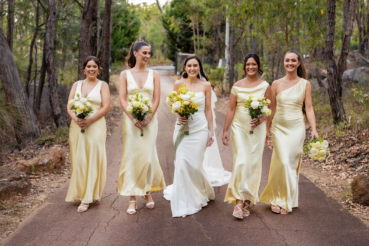 Bride walks with four bridesmaids in matching yellow satin dresses carrying bouquets along a forest path.