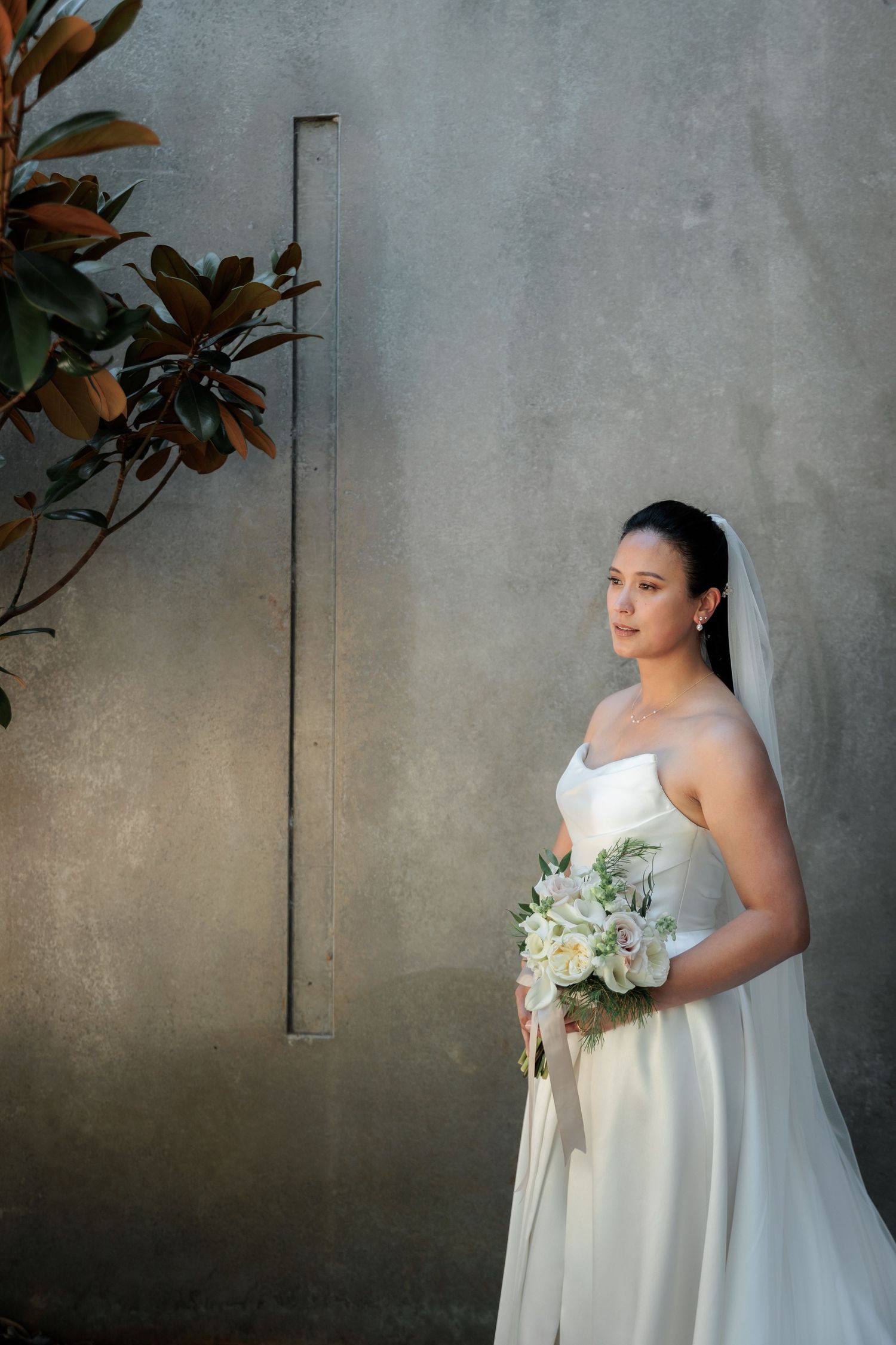 Elegant bride in strapless white gown holding floral bouquet, standing against concrete wall with veil and earrings.