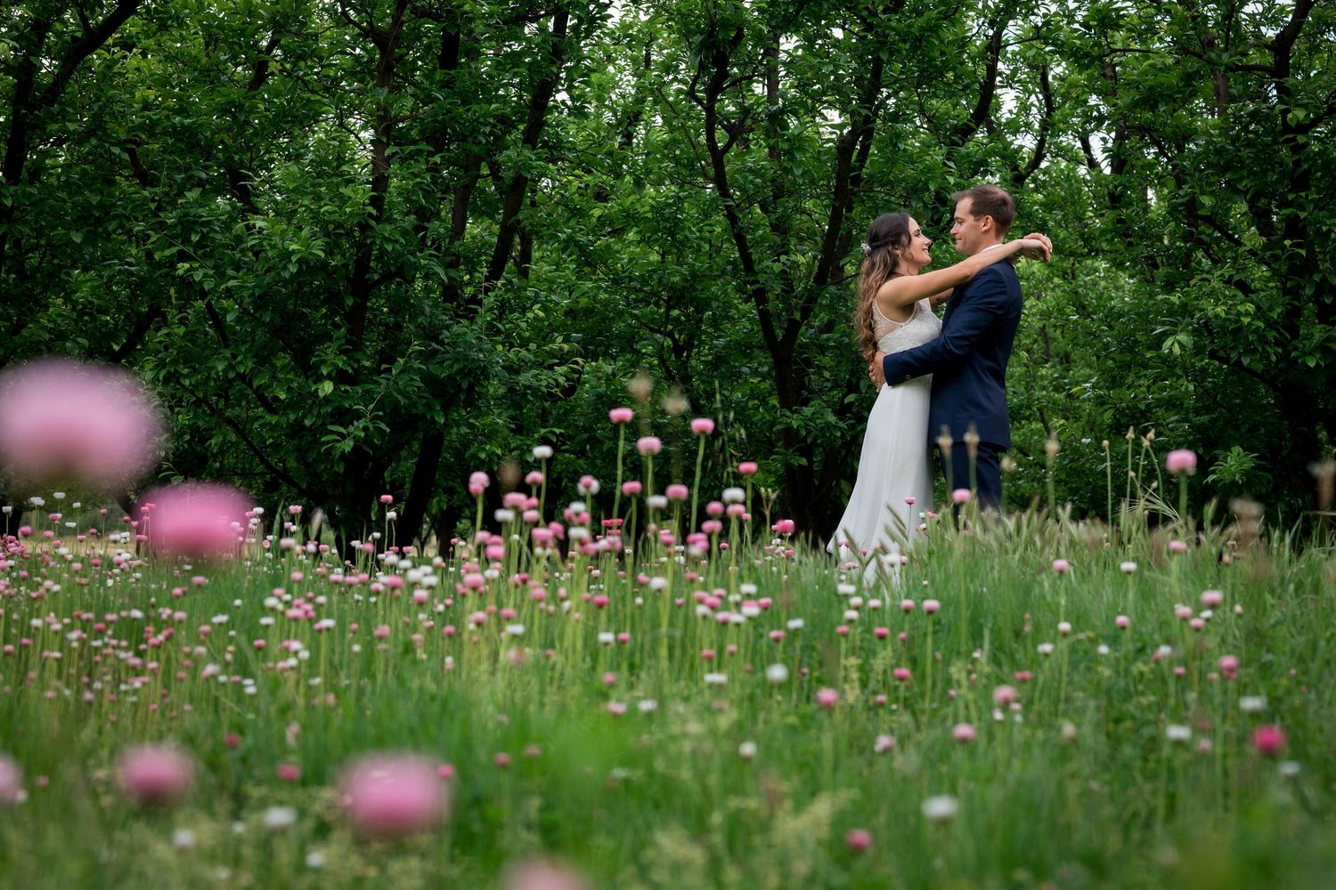 Backyard wedding - Bride and groom embracing in a lush green field of pink wildflowers during an outdoor wedding ceremony.