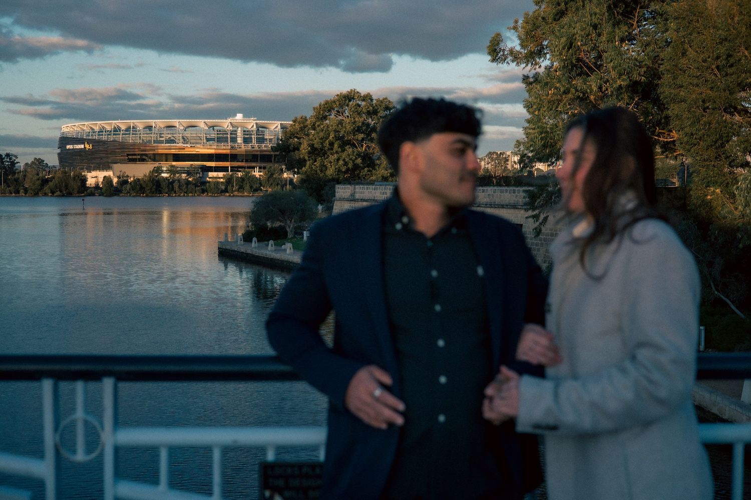 Two people share a tender moment by the waterfront during sunset with city buildings in the background.