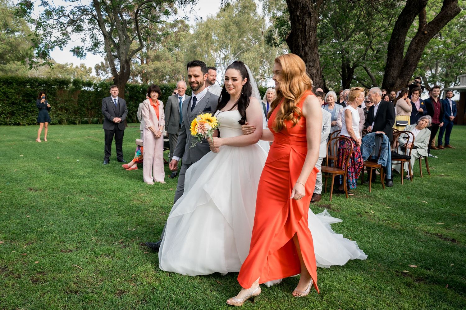 Outdoor wedding ceremony with guests seated on lawn under mature trees.