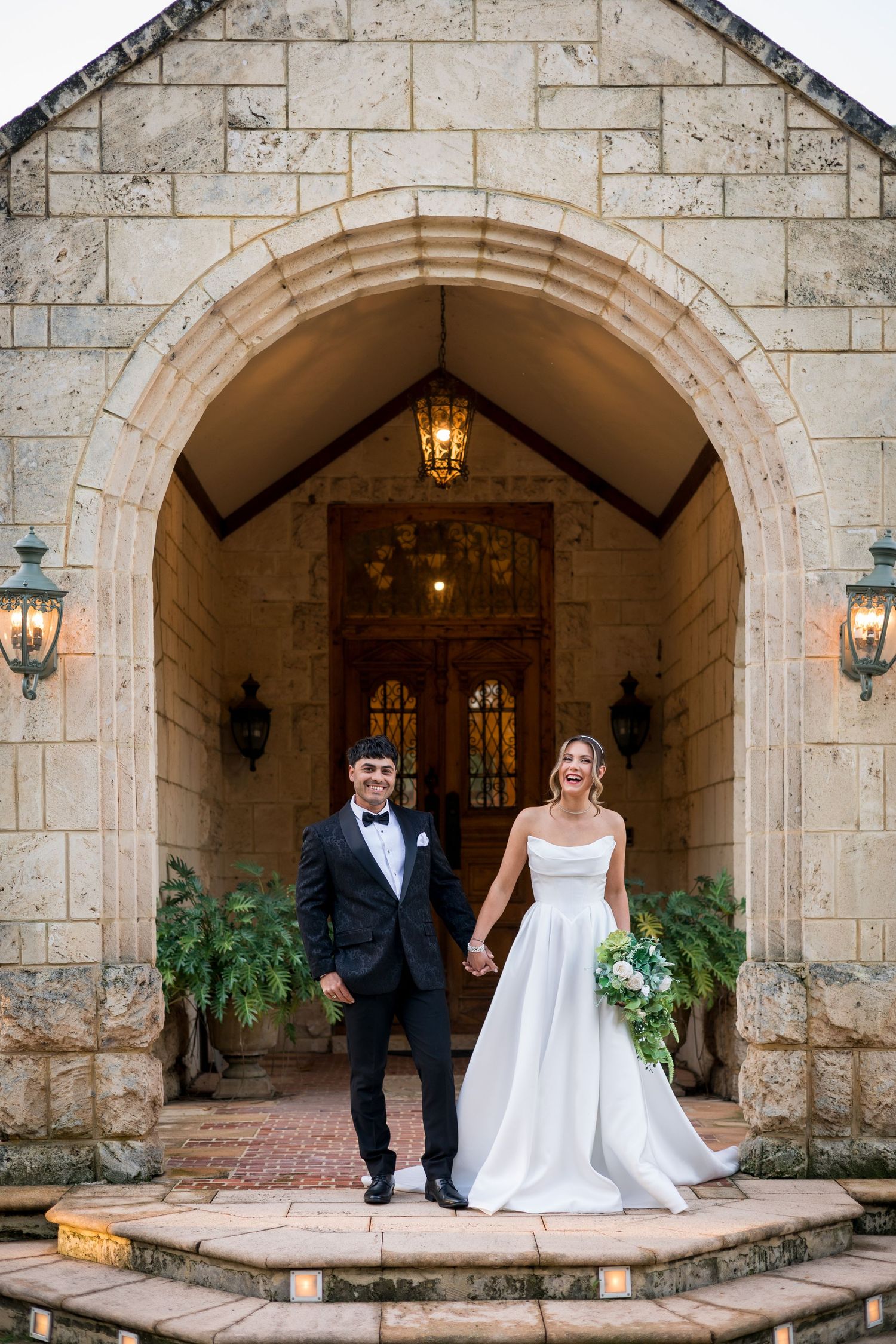 Bride and groom holding hands under stone arch at elegant wedding venue with warm lighting and wooden doors.