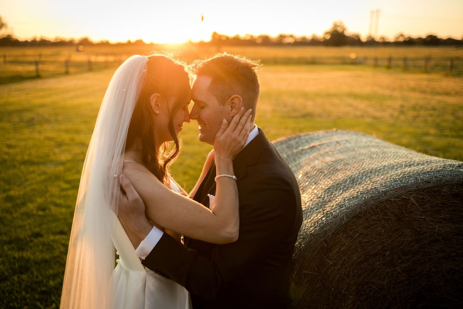 Bride and groom share romantic golden hour moment beside hay bale in sunlit field, bride in white veil, groom in black suit.